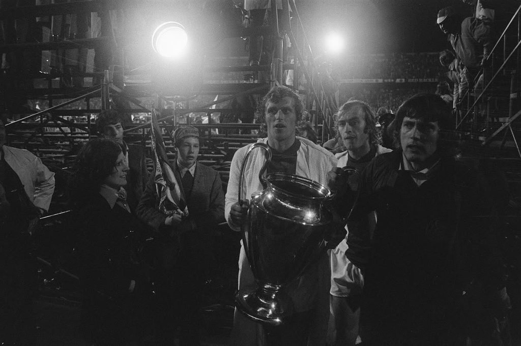 Ajax players carrying the European Cup trophy under the floodlights, with fans behind them.