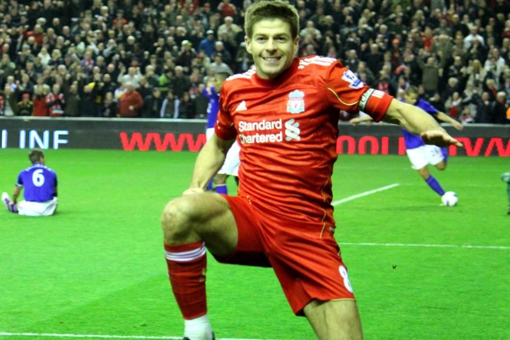 Steven Gerrard in a red Liverpool shirt celebrating on one knee in front of the Anfield crowd.