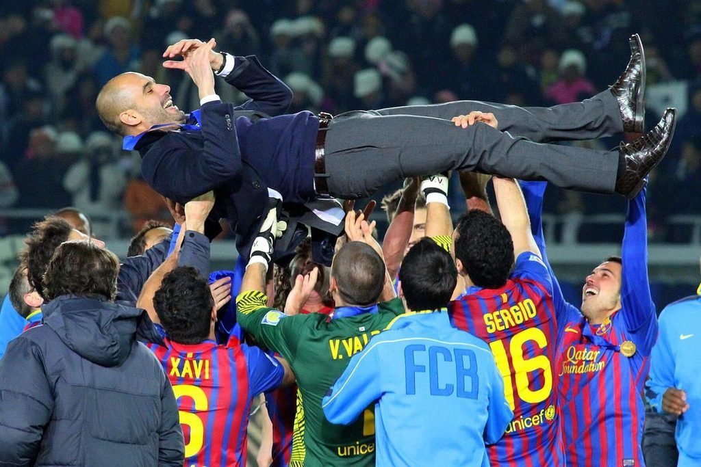 Barcelona players in red and blue shirts lifting Pep Guardiola, in a suit, above their heads.