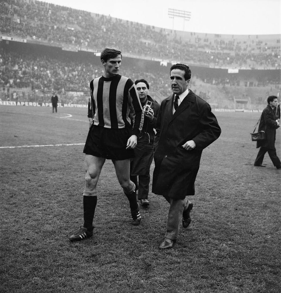 An Inter player in a striped shirt walking across the San Siro pitch beside Helenio Herrera in a long dark coat.