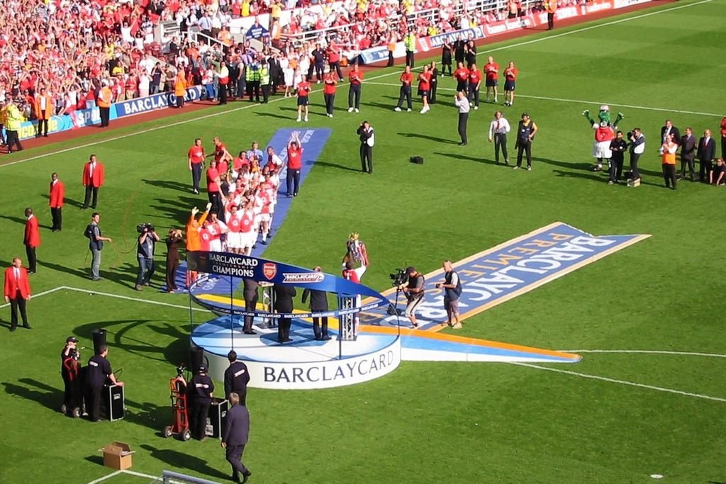 Arsenal players on a stage on the Highbury pitch with the Premier League trophy, in front of a full crowd.