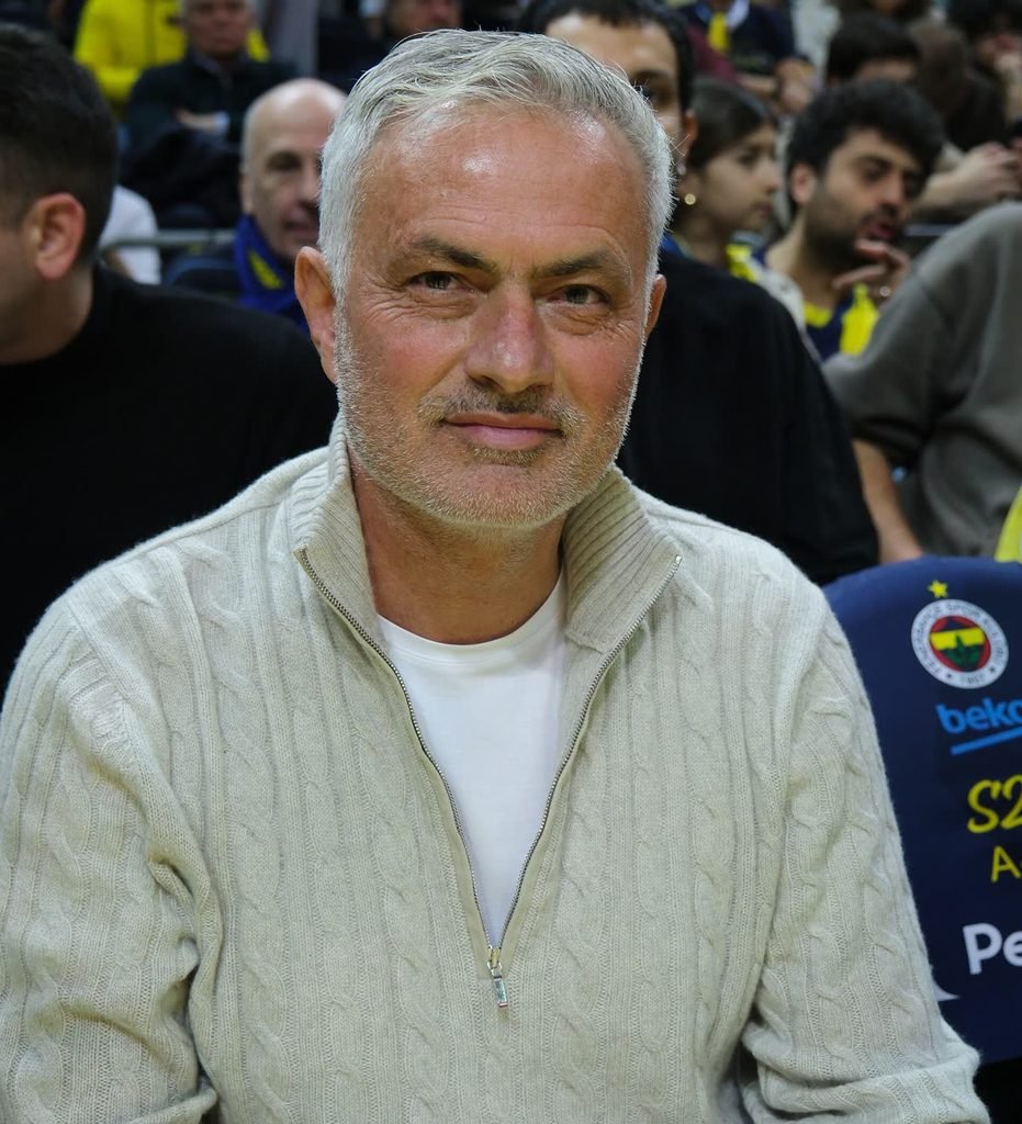 José Mourinho, grey-haired, smiling slightly in a light knitted jumper, with a crowd behind him.