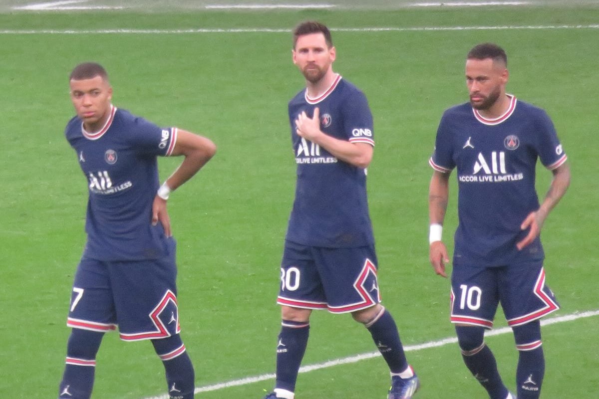 Kylian Mbappé, Lionel Messi and Neymar walking side by side on the pitch in dark blue PSG shirts.
