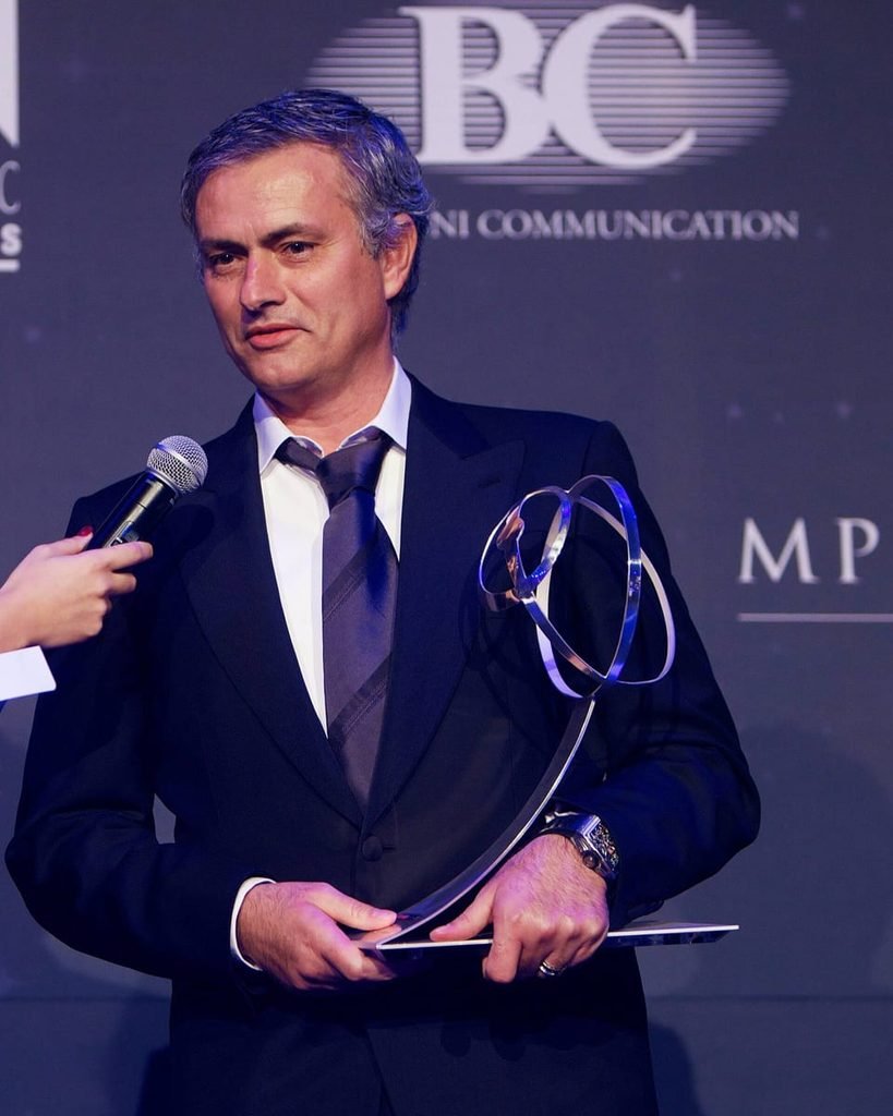 José Mourinho in a dark suit, smiling slightly, holding a glass trophy at an awards ceremony.
