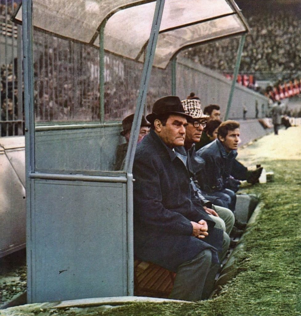 An older man in a dark coat and hat sitting on a covered dugout bench beside the pitch, a full stand behind him
