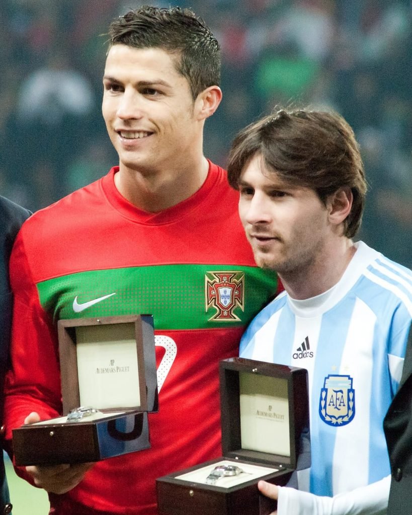 Cristiano Ronaldo in a red Portugal shirt and Lionel Messi in Argentina blue and white, side by side, each holding a watch box.
