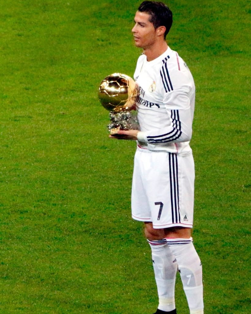 Cristiano Ronaldo standing on the pitch in a white Real Madrid kit, holding the golden Ballon d'Or trophy.